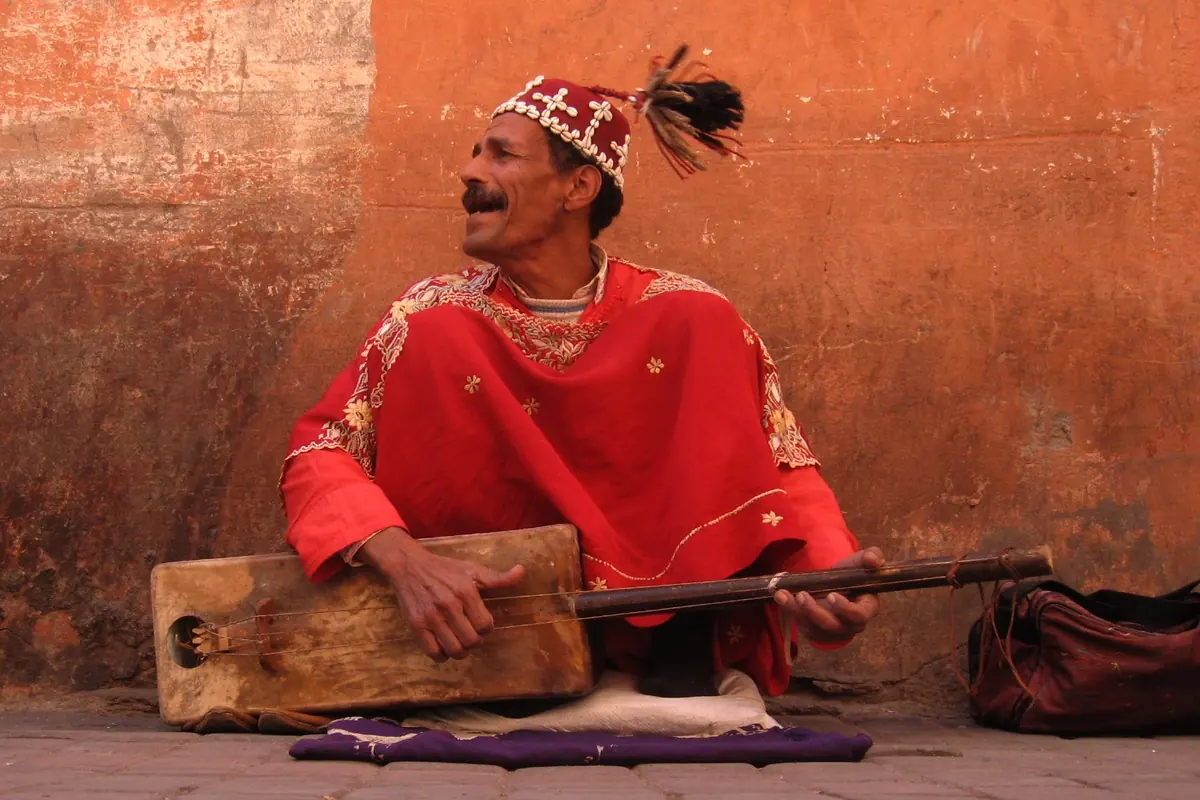 A guimbri player in a red robe and a fringed cap sits against an orange wall in the Marrakech medina